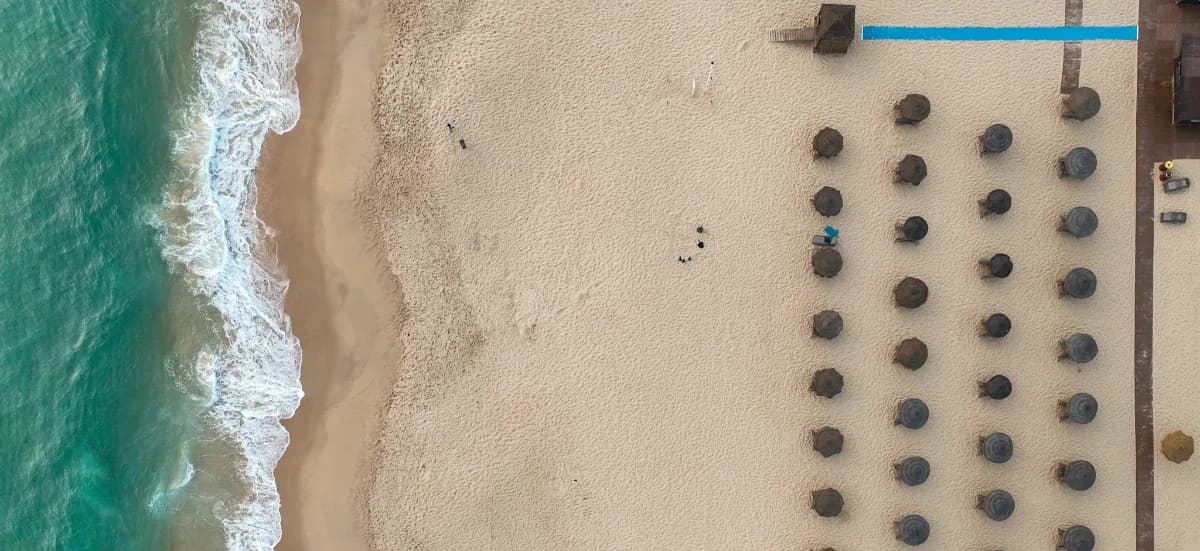 Aerial view of a beachfront hotel — rows of umbrellas on white sand at the water's edge